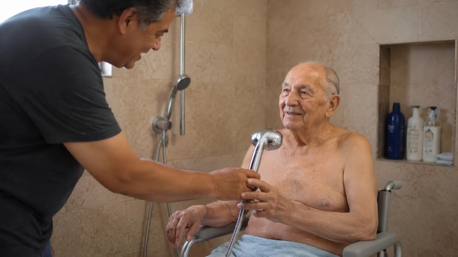 Elderly man sitting comfortably on a padded shower chair while a caregiver hands him a handheld shower head in a walk-in shower