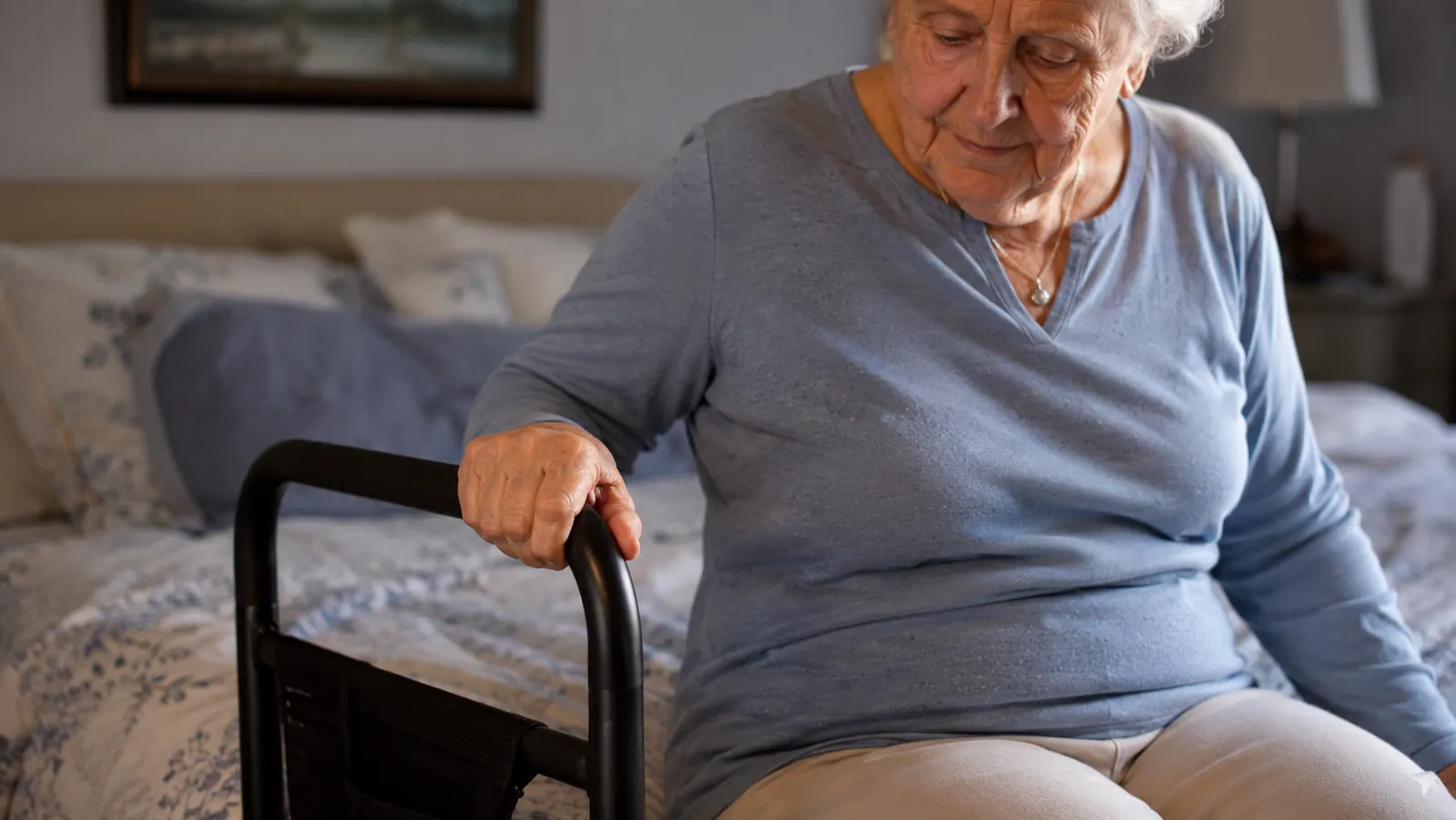 Elderly woman using a bed assist bar to push herself upright from a low mattress in a warmly lit bedroom
