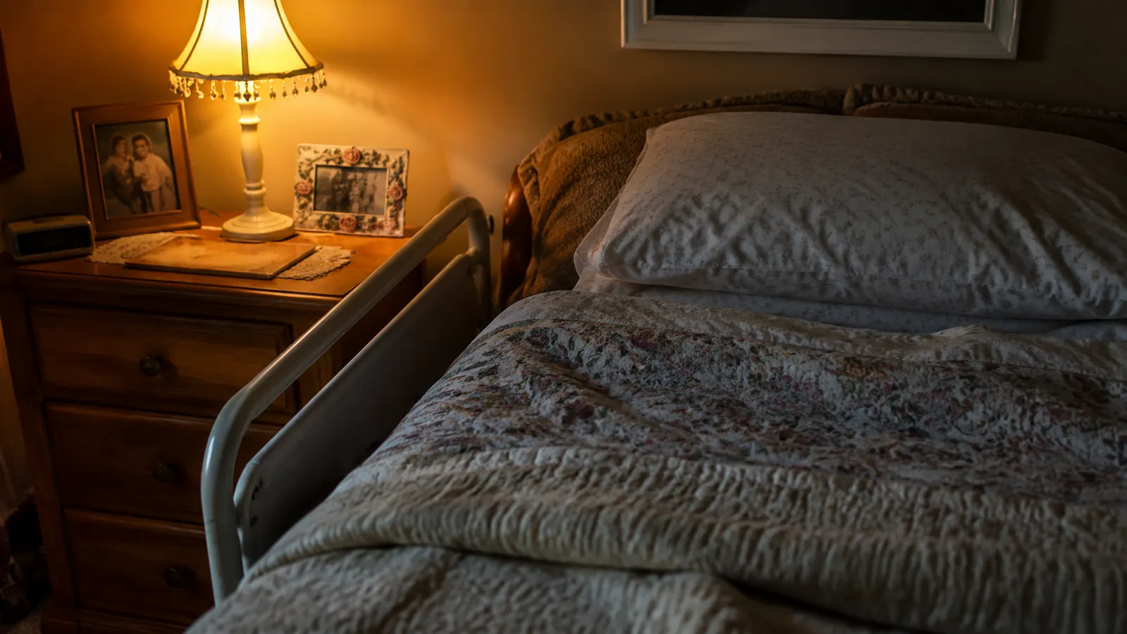 Soft bedroom scene at night, warm lamp light on a neatly made bed with a small rail visible at the side, calm and reassuring